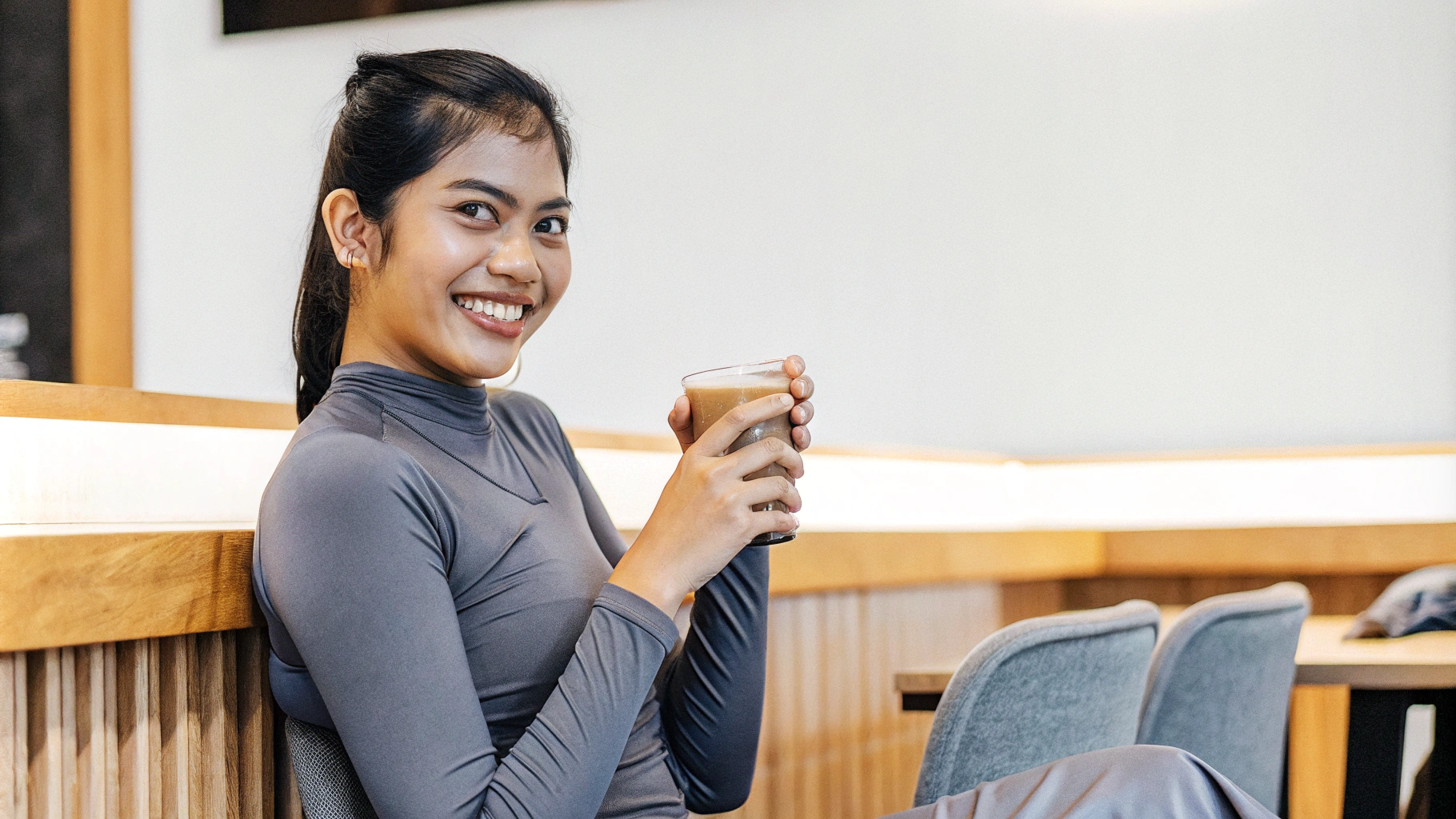 AI generated image by Red Panda AI: a beautiful Indonesian woman in a tight fitness t-shirt, full body, sitting in a cafe smiling looking at the camera while holding a clear glass cup containing brown late coffee